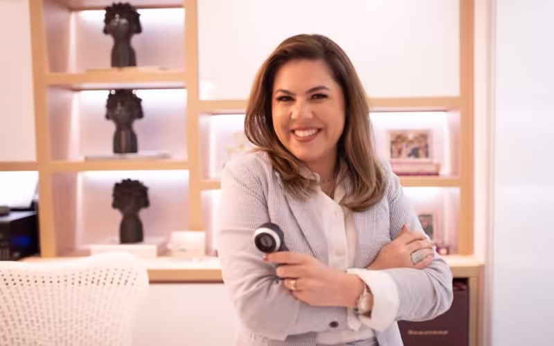 Dra. Aline Soares de braços cruzados, em pé diante das prateleiras iluminadas da sala de espera da clínica.