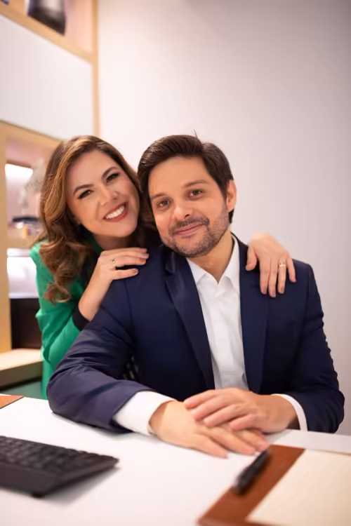Dra. Aline Soares, de blazer verde, apoiada no ombro do Dr. Osório Lara, sentado à mesa de atendimento da clínica.