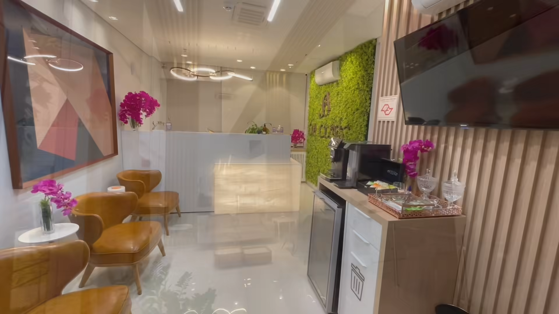 Recepção da OA Clinic vista da sala de espera, com poltronas de couro caramelo, orquídeas e a parede de vegetação ao fundo.